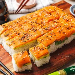 Rechteckiges Sushi mit Reis, Avocado und orangefarbener veganer Sushi Füllung, bestreut mit Sesam. Sojasauce und Wasabi sind daneben.