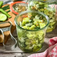 Frischer gurkensalat ohne zucker, zubereitet mit Dill und Senfkörnern, in Gläsern abgefüllt. Gurken und Sparschäler liegen bereit.