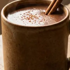 Eine dampfende Tasse vegane heiße Schokolade mit Zimtstange und Kakaopulver auf einem Holztisch.