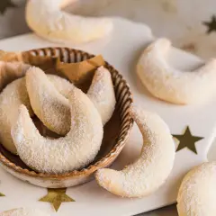 Vegane Vanillekipferl rezept: zarte, mondförmige Kekse, bestäubt mit Puderzucker, liegen in einer Schale und auf einem Brett, umgeben von goldenen Sternen.