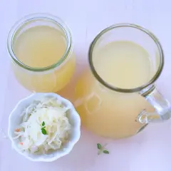 Ein Glas und ein Krug mit Sauerkrautsaft, reich an Vitaminen, stehen neben einer Schale Sauerkraut.