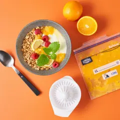 Orangensaft in einem Gefrierbeutel zeigt, dass man orangen einfrieren kann. Daneben ein Müsli mit Früchten.