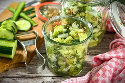 Frischer gurkensalat ohne zucker, zubereitet mit Dill und Senfkörnern, in Gläsern abgefüllt. Gurken und Sparschäler liegen bereit.
