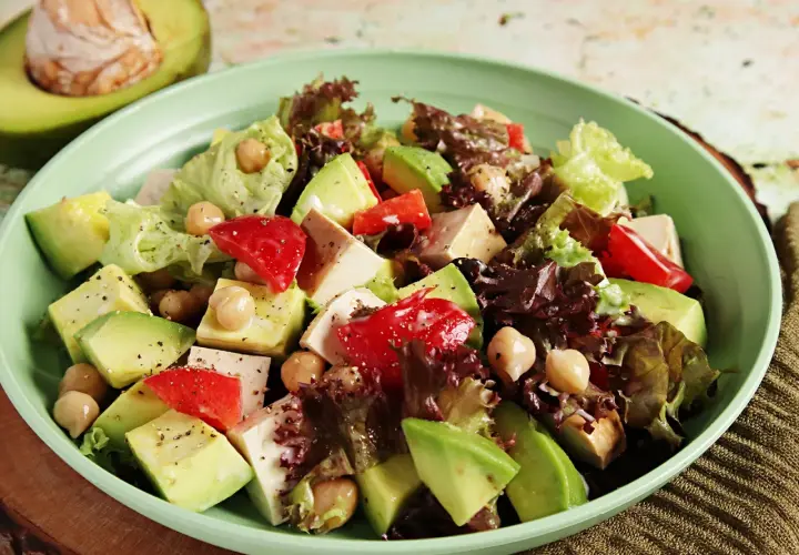 Frischer veganer Salat mit Avocado, Kichererbsen, Tofu und roten Paprikastücken in einer hellgrünen Schüssel.