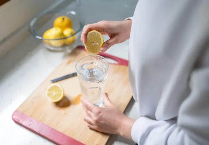 Eine Hand presst eine Zitronenhälfte über ein Glas Wasser. So beginnt der Tag mit erfrischendem Zitronenwasser.