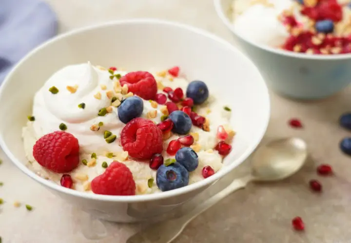 Veganes Grießbrei-Dessert mit frischen Himbeeren, Blaubeeren, Granatapfelkernen und gehackten Nüssen, garniert mit einem Klecks veganer Sahne.