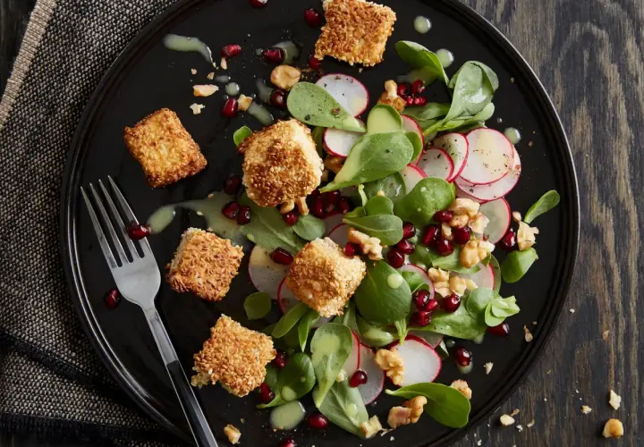 Feldsalat mit Granatapfelkernen, Radieschen und Sesam-Käsewürfeln auf schwarzer Platte.