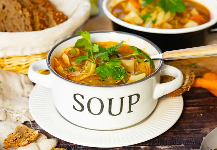 Herzhafte Kohlsuppe, low carb, in einem weißen Topf mit der Aufschrift "SOUP", garniert mit Petersilie. Dazu gibt es Vollkornbrot und Karotten.