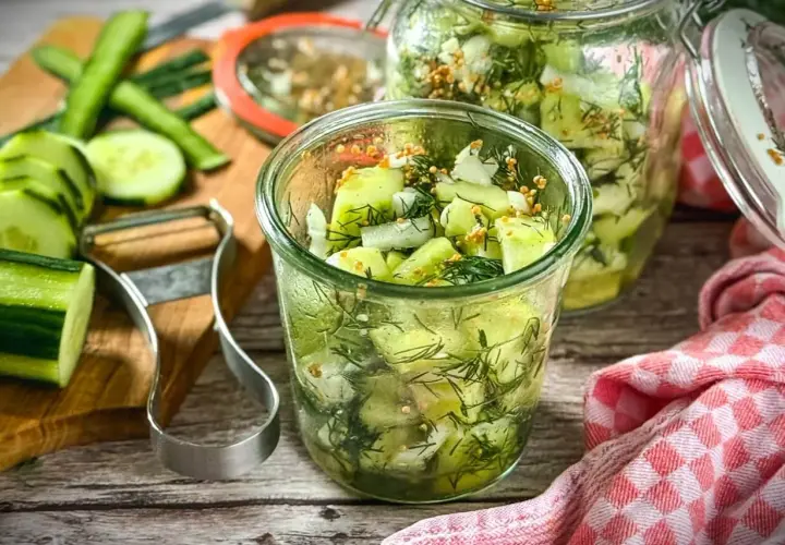 Frischer gurkensalat ohne zucker, zubereitet mit Dill und Senfkörnern, in Gläsern abgefüllt. Gurken und Sparschäler liegen bereit.
