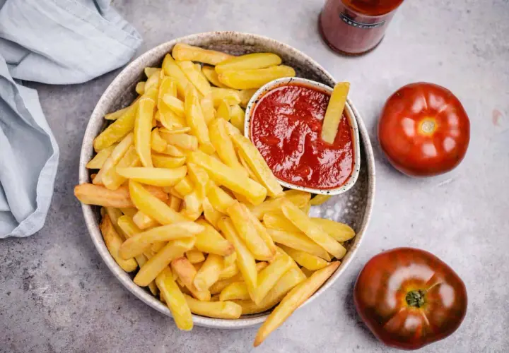 Knusprige Pommes Frites mit einem Schälchen veganem Ketchup. Zwei Tomaten und eine Flasche Ketchup liegen daneben.