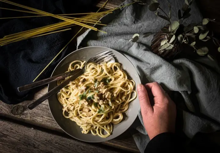 Teller mit cremiger, veganer Spaghetti Carbonara, garniert mit Kräutern. Daneben liegen ungekochte Spaghetti.