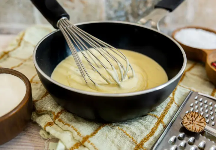Eine cremige low carb bechamel wird in einer Pfanne mit einem Schneebesen verrührt. Daneben stehen Schüsseln mit Sahne, Salz und Pfefferkörnern.