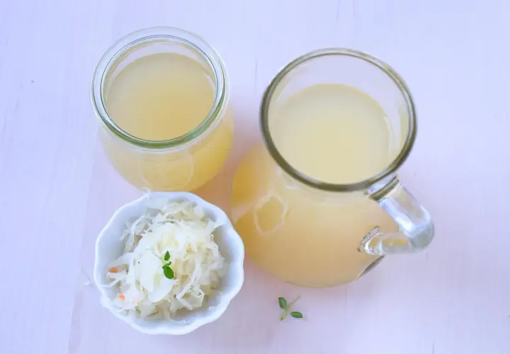 Ein Glas und ein Krug mit Sauerkrautsaft, reich an Vitaminen, stehen neben einer Schale Sauerkraut.
