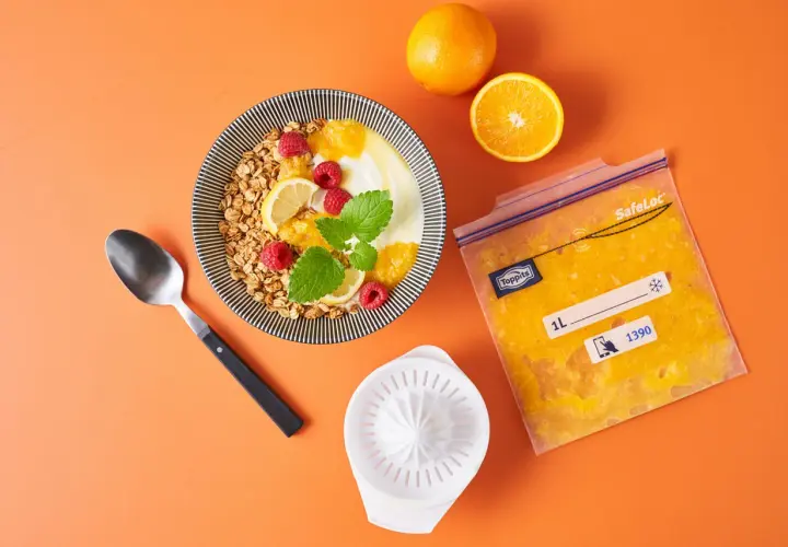 Orangensaft in einem Gefrierbeutel zeigt, dass man orangen einfrieren kann. Daneben ein Müsli mit Früchten.