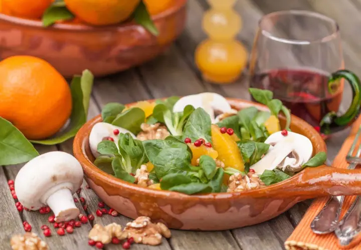 Ein frischer Wintersalat mit Orangen, Pilzen und Walnüssen in einer Terrakotta-Schale. Daneben liegen frische Orangen und ein Glas Rotwein.