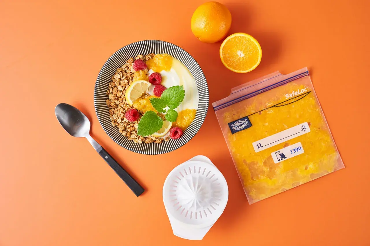 Orangensaft in einem Gefrierbeutel zeigt, dass man orangen einfrieren kann. Daneben ein Müsli mit Früchten.