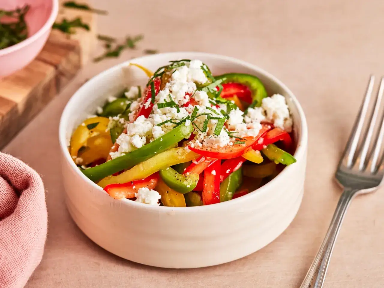 Bunter Paprika-Salat mit Feta-Käse und Kräutern in einer Schüssel, daneben eine Gabel und ein rosa Tuch.