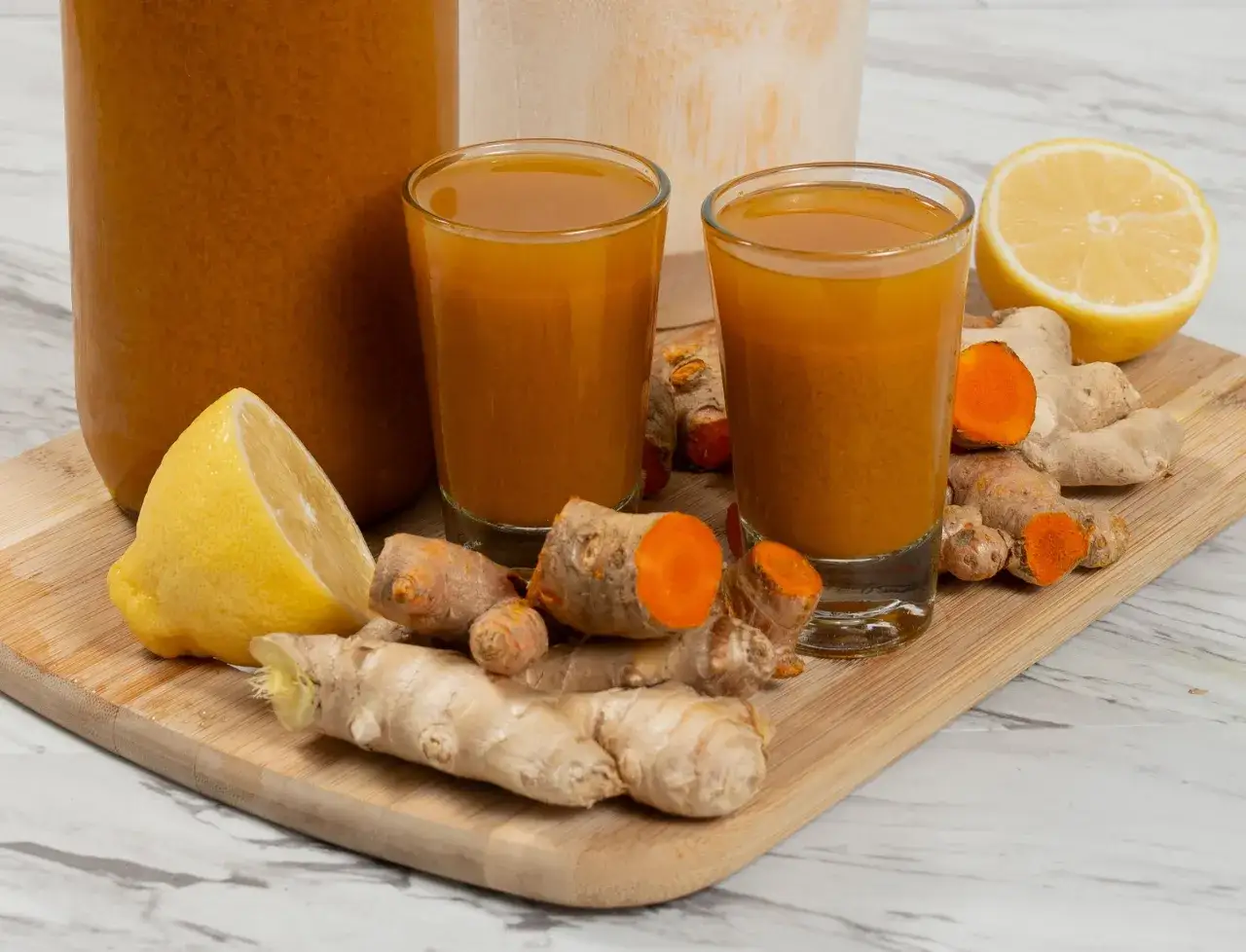 Zwei Gläser mit leuchtend gelbem Ingwer Kurkuma Shot, daneben frische Ingwerwurzeln, Kurkuma und Zitronenspalten auf einem Holzbrett.
