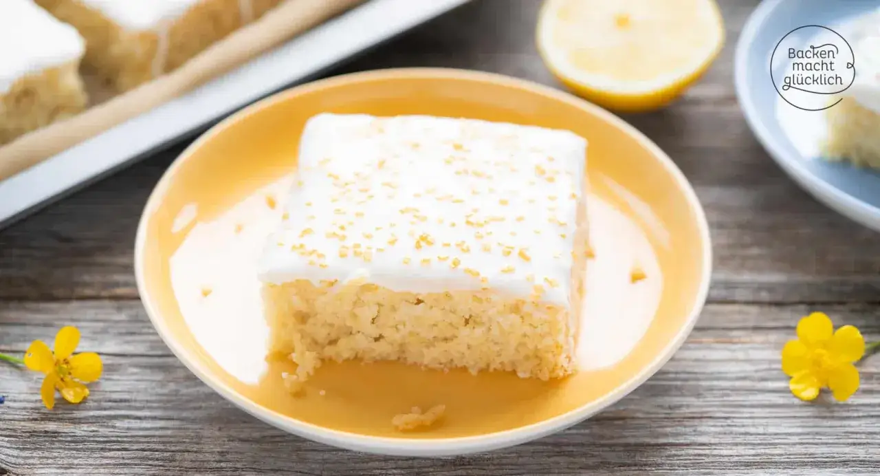 Ein Stück saftiger veganer Blechkuchen mit weißer Glasur und goldenen Streuseln auf einem gelben Teller, daneben eine Zitronenspalte.